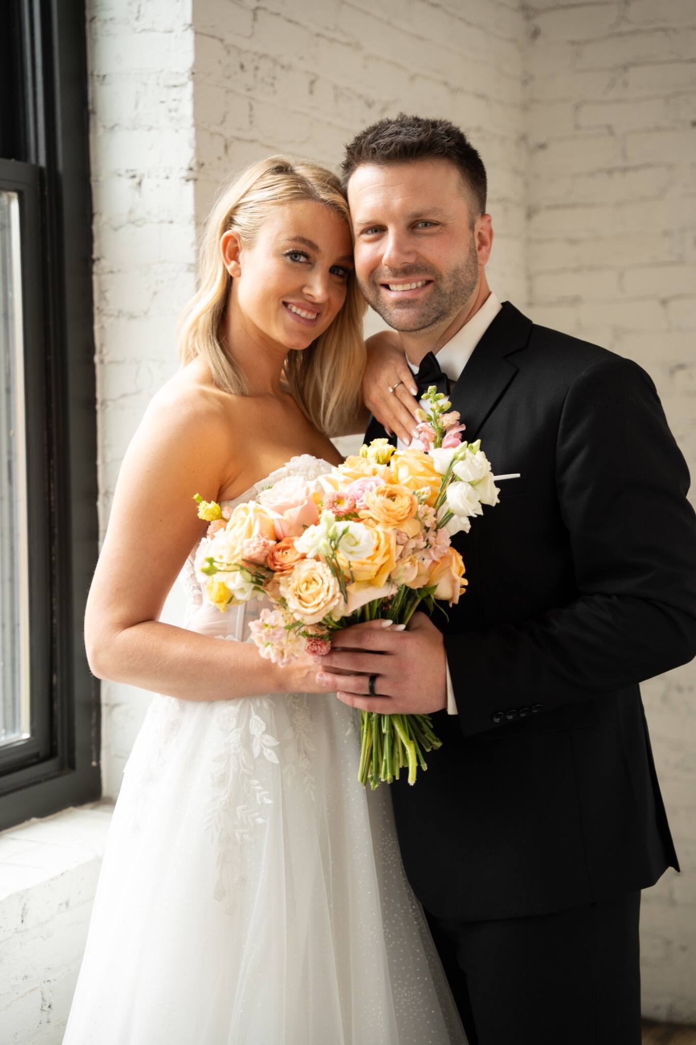 Newlyweds embracing by a window with wedding flowers