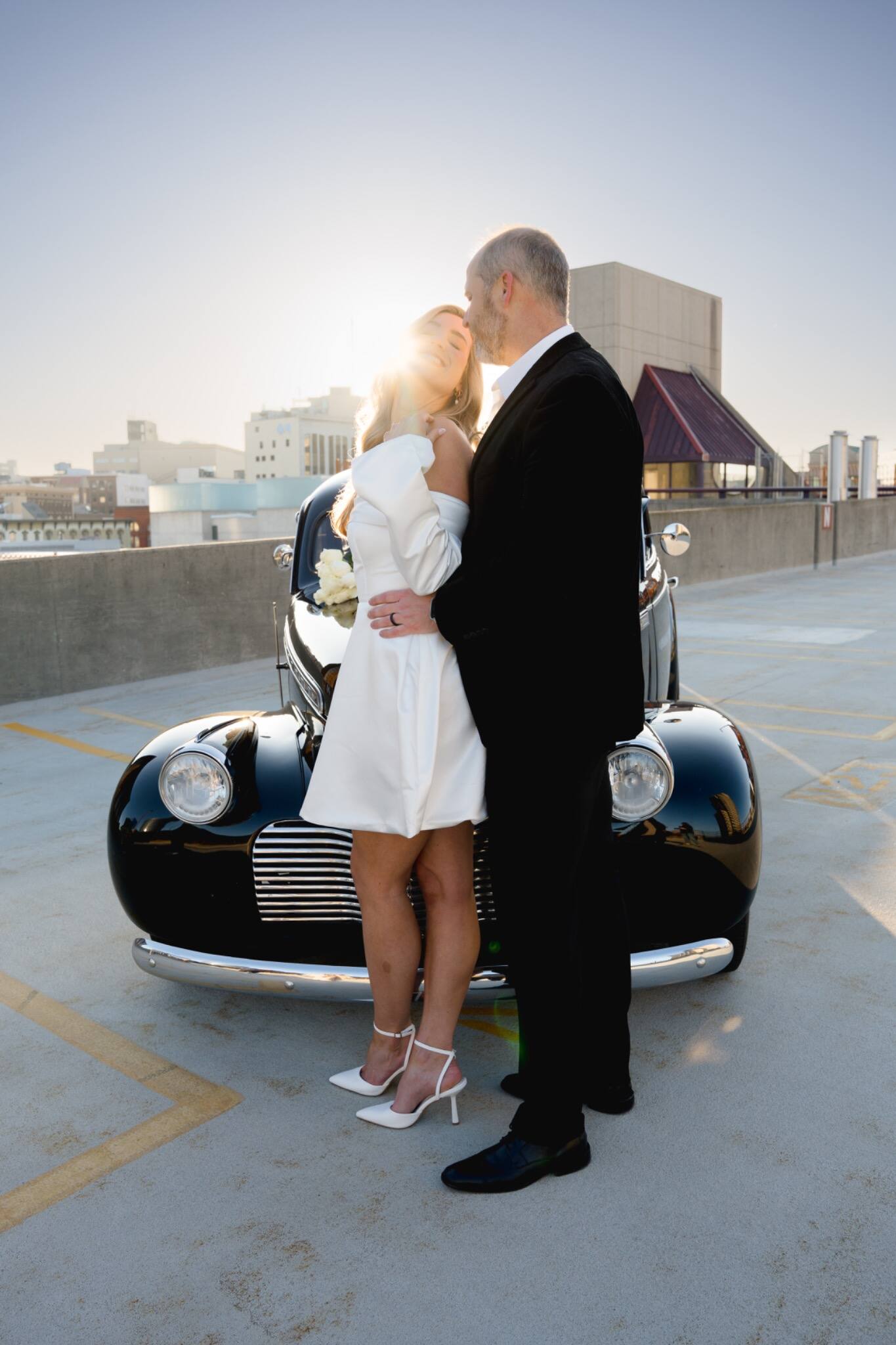 A newlywed couple kissing beside their vintage car